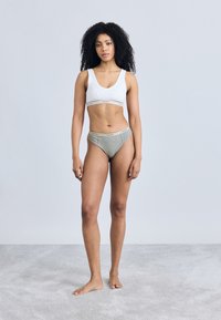 Λευκό μπραλέτ με φαρδιά ταινία, σετ με γκρι hipster σλιπ με αντίθετη ζώνη μέσης. Το μοντέλο στέκεται ξυπόλητο σε ένα ανοιχτό χαλί.