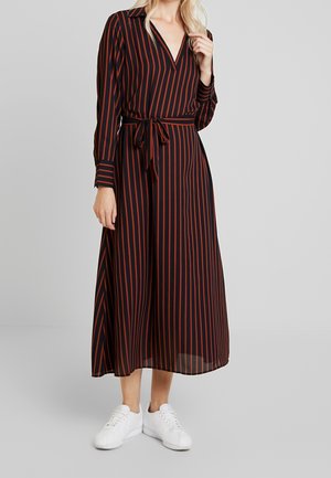 Femme portant une robe midi noire à rayures rouges verticales, manches longues, col, ceinture nouée à la taille, et baskets blanches, debout sur un sol gris.