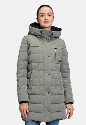 Vrouw draagt een grijze gewatteerde winterjas met capuchon, ritssluiting en drukknopen, staand tegen een effen lichte achtergrond.