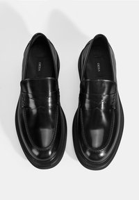 Ein Paar polierte schwarze Leder-slipper mit abgerundeten Zehen und dicken Gummisohlen, Markenname "DESA" innen sichtbar.