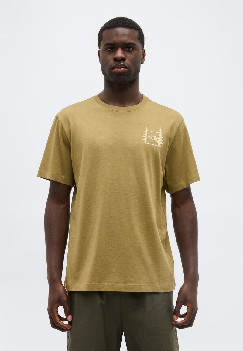 Mann trägt ein olivgrünes T-Shirt mit einem kleinen Wald-Logo auf der Brust, steht vor einem schlichten hellen Hintergrund und blickt nach vorne.