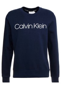 Granatowa bluza Calvin Klein z okrągłym dekoltem, długimi rękawami i wyraźnym białym logo nadrukowanym na przodzie. Miękka tekstura materiału.