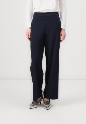 Pantalons larges bleu marine avec une texture lisse, taille haute et devant plissé, associés à des chaussures pointues métalliques.
