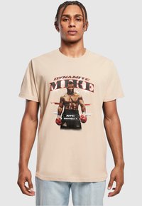 Tricou din bumbac bej, prezentând o imprimare grafică cu un boxer purtând mănuși roșii, textul „DYNAMITE MIKE” și „NYC BROOKLYN” în litere îngroșate.