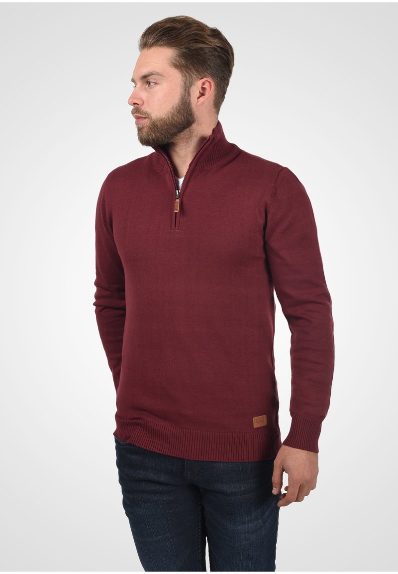 Maglioncino bordeaux con zip e collo alto, tessuto a maglia, polsi e orlo rigati, con una piccola etichetta in pelle nell'angolo in basso a sinistra.