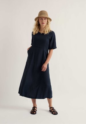 Žena s blond vlasmi, ktorá má na sebe opálený pletený klobúk, námornícky modré midi šaty s krátkymi rukávmi a vreckami a čierne sandále na remienky, stojaca pred jednoduchým pozadím.