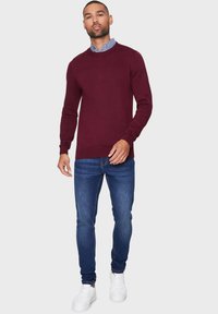 Maglione a coste color borgogna con scollo rotondo, abbinato a jeans blue slim-fit e sneaker bianche, con una texture morbida e un design aderente.