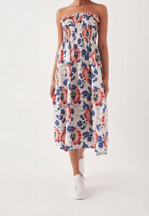 Strapless midi-jurk met rood-blauw bloemenpatroon, een gerimpeld lijfje, gedragen met witte sneakers, model loopt naar voren op een effen achtergrond.