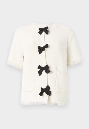 Short-sleeve cream sweater with a textured knit, featuring four black bow closures, two front pockets, and fringed hem details.