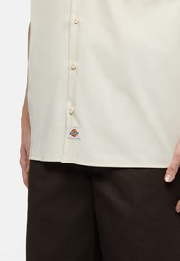 Camicetta crema a maniche corte con bottoni, dalla texture liscia, con un piccolo etichetta del logo Dickies all'orlo, abbinata a pantaloni marroni scuri.