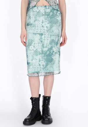 Femeie purtând o fustă verde până la mijlocul gambelor cu model patchwork, asortată cu bocanci negri cu șiret, stând în fața unui fundal alb.