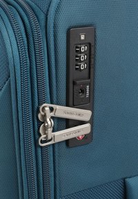 Valigia di tessuto color teal con doppi zip, dotata di lucchetto a combinazione approvato dalla TSA e di una toppa di sicurezza rettangolare. Texture liscia, design resistente.