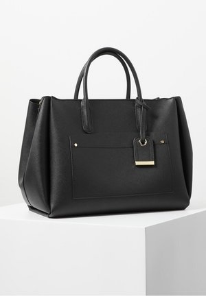 Czarna skórzana torebka z podwójnymi uchwytami, przednią kieszenią oraz dołączoną zawieszką ze złotym obszyciem, prezentowana na białym piedestale na jednolitym tle.