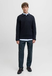 Dunkelblauer Strickpullover mit kragenförmigem Ausschnitt und drei Knöpfen, kombiniert mit dunkelgrünen Hosen und braunen Schuhen.