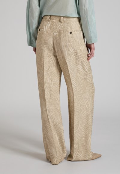 Pantaloni beige a gamba larga con motivi di foglie in rilievo, tasche posteriori con bottoni, indossati con una maglia a maniche lunghe azzurro chiaro e scarpe beige con punta.