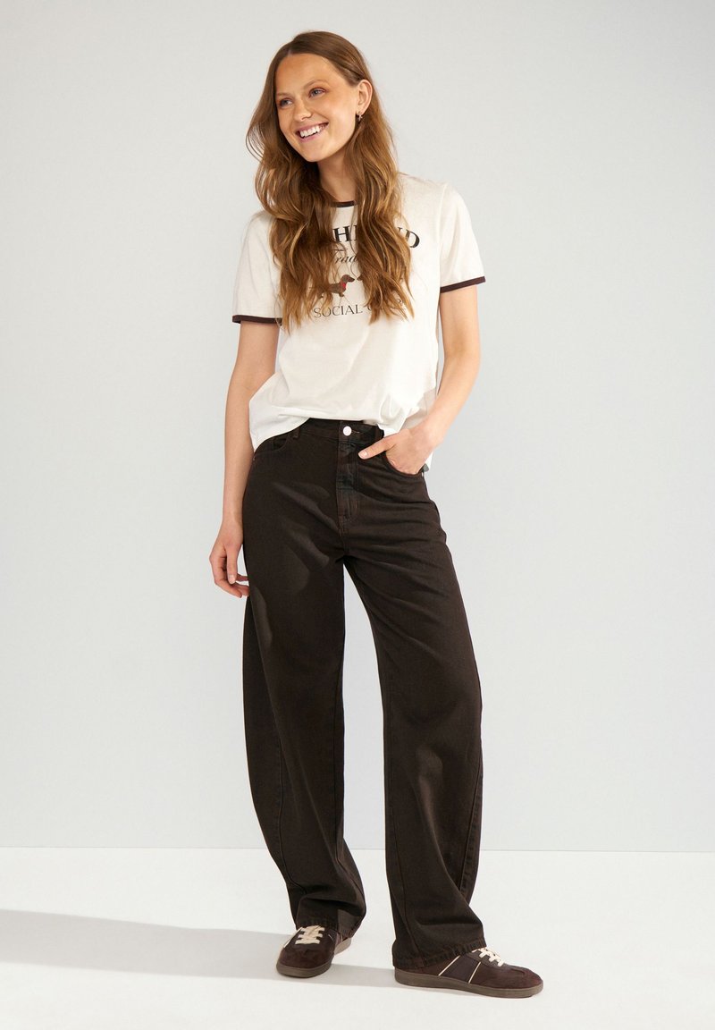 Femme aux longs cheveux portant un t-shirt graphique blanc, un pantalon noir à jambes larges et des baskets marron, debout devant un fond uni.