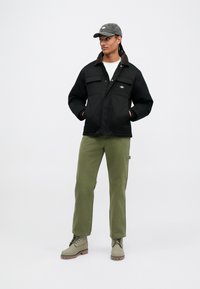 Homem vestido com um casaco preto, uma camisa branca, calças verde-oliva, botas cinzas e um boné escuro, de pé com as mãos nos bolsos do casaco.