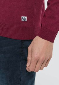 Pull en tricot bordeaux avec des poignets et un ourlet côtelés. Présente une petite étiquette de marque blanche sur le côté inférieur. Associé à un jean en denim foncé.
