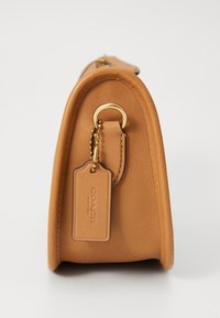 Geantă de umăr din piele maro deschis, cu o formă rotundă și accesorii aurii, având un etichetă suspendată prinsă cu o lanț. Textură netedă și design minimal.