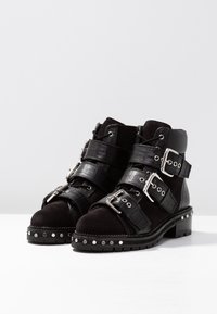 Botines negros de ante y cuero texturizado, con tres correas ajustables, hebillas de metal y detalles de tachuelas plateadas en la suela.