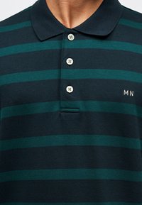 Homme portant un polo rayé vert foncé et noir, avec trois boutons blancs et une petite broderie "MN" sur la poitrine.