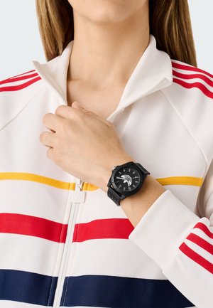 Montre noire avec un cadran rond, des marquages blancs et un bracelet en silicone noir. Portée sur une main au-dessus d'une veste blanche à rayures multicolores.