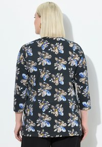 Frau mit blonden Haaren, die ein schwarzes Oberteil mit blau-beigen Blumenmuster trägt, von hinten vor einem weißen Hintergrund gezeigt.
