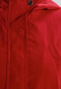 Röd kappa med huva, mjuk textur, dragkedjestängning och en tryckknapp som accent. Innehåller en sidoficka och synliga sömmar.