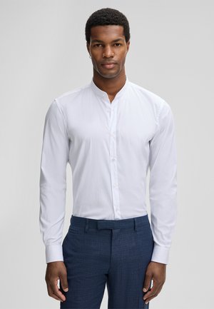 Camicia bianca a maniche lunghe con colletto a banda, caratterizzata da una superficie liscia e un design aderente. Indossata con pantaloni blu scuro texturizzati.