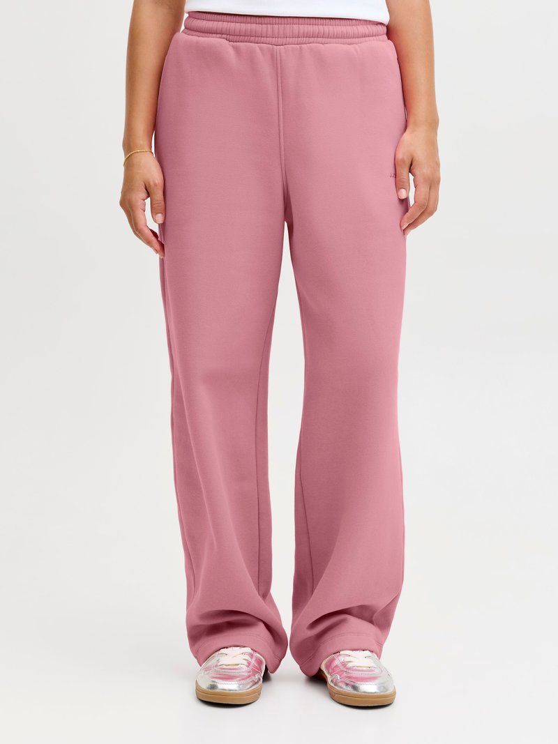Vrouw draagt losse roze sweatpants en zilverroze sneakers, staand tegen een effen witte achtergrond.