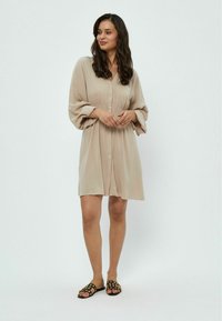 Abito beige con maniche lunghe e ampie, vita arricciata, caratterizzato da una chiusura con bottoni e una silhouette fluida. Abbinato a sandali neri.