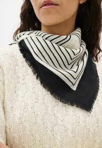 Kvinde iført et sort og hvidt stribet trekantet tørklæde med frynser over en cremet kabelstrik sweater.