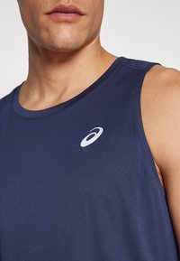 Navy blue sleeveless athletic shirt made of fabric with a smooth texture, featuring a white logo on the chest. Close-up of neckline and shoulder.