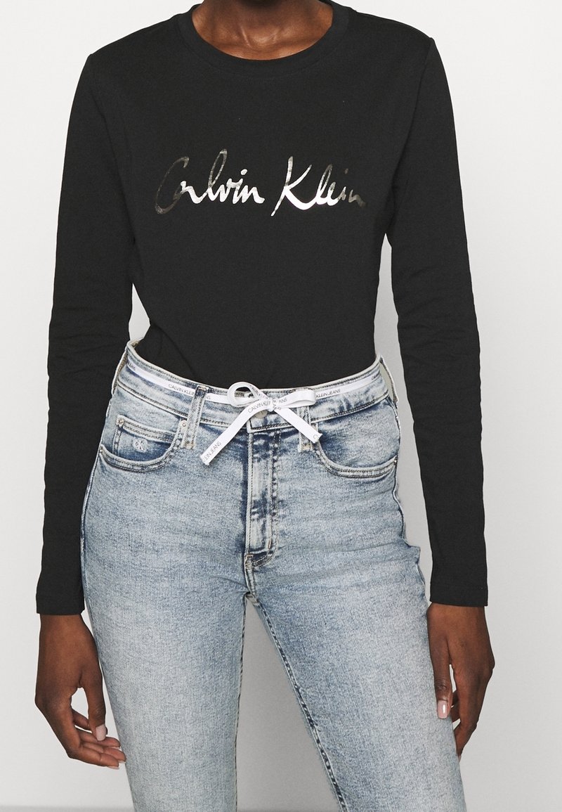 Svart långärmad T-shirt med "Calvin Klein" i metallisk bokstäver, ihop med ljusblå högmidjade jeans med vit dragsko.
