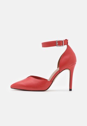 Czerwona szpilka z wyciągniętym noskiem i paskiem wokół kostki, z wykończeniem w postaci srebrnej klamry, przedstawiona w profilu bocznym na białym tle.