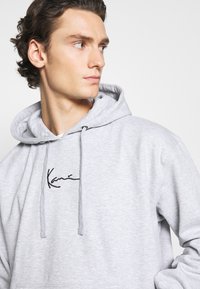 Grå sweatshirt med hætte, prydet med en sort broderet signatur på brystet. Lavet af blødt, tekstureret materiale med snoretræksdetaljer.