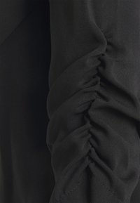 Lähikuva mustasta kankaasta, jossa on poimutettua yksityiskohtaa, joka luo materiaalin pinnalle kerääntyneitä, teksturoiduista laskoksia.