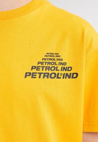 Camiseta de algodón amarilla con texto repetido en azul oscuro "PETROL IND" en diferentes tamaños en la zona del pecho izquierdo. Diseño clásico de cuello redondo.