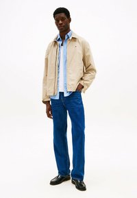 Beige lichtgewicht jas met een kraag, gedragen over een blauw-wit gestreept shirt, gecombineerd met blauwe jeans en zwarte schoenen. Eenvoudig, schoon ontwerp.