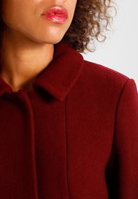 Cappotto in lana borgogna con colletto arrotondato e tessuto a trama intricata, caratterizzato da un dettaglio di bottone chiuso all'ascella.