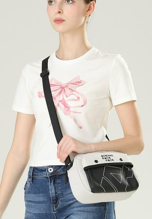 Giovane donna con maglietta bianca con stampa di fiocco rosa, jeans blu e una borsa a tracolla bianca e nera con testo sulla patta.