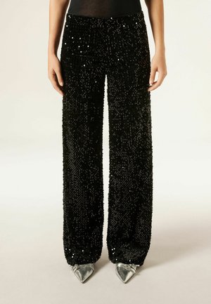 Černé široké kalhoty se flitry a texturovaným povrchem, s vysokým pasem a hladkým povrchem. Stříbrné boty s špičatou špičkou doplňují vzhled.