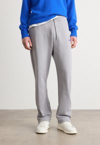 Pantalones de chándal gris claro con cintura elástica, cordón y bolsillos laterales, combinados con un suéter azul y zapatillas blancas.