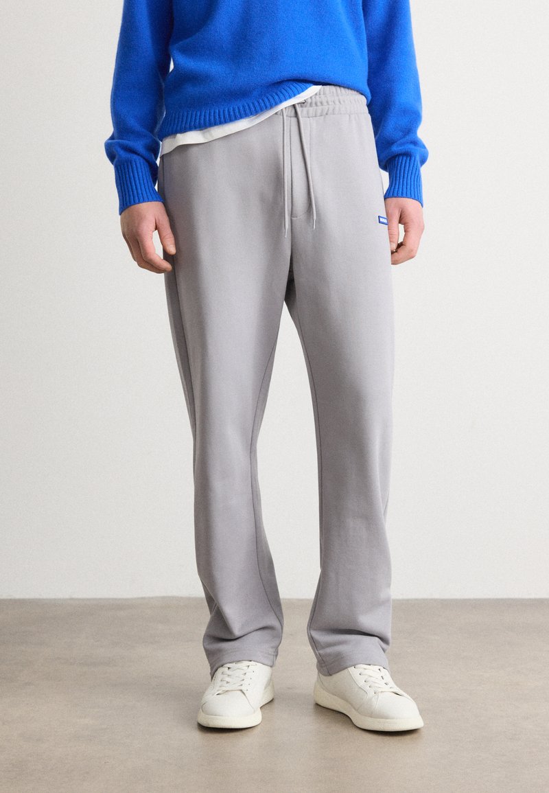 Pantalones de chándal gris claro con cintura elástica, cordón y bolsillos laterales, combinados con un suéter azul y zapatillas blancas.
