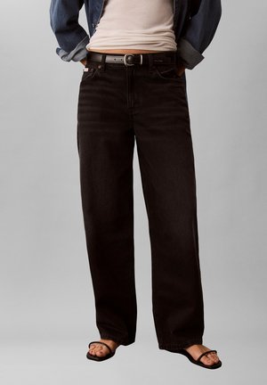 Person iført løse sorte jeans med sort bælte, en hvid skjorte, blå denimjakke og sorte sandaler med stropper.
