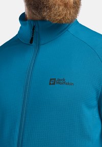 Veste zippée turquoise en tissu texturé, dotée d'un col montant et d'un logo noir discret sur le côté gauche.