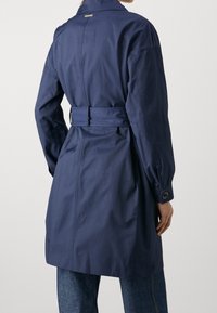 Granatowy płaszcz trencz z paskiem w talii, długimi rękawami i zapinanymi mankietami; posiada wyraźny kołnierz i gładką fakturę materiału.