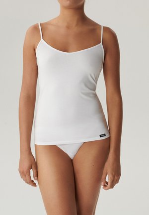 Kobieta ubrana w białą koszulkę na cienkich ramiączkach oraz dopasowane białe figi, stojąca na tle jasnego, jednolitego tła.