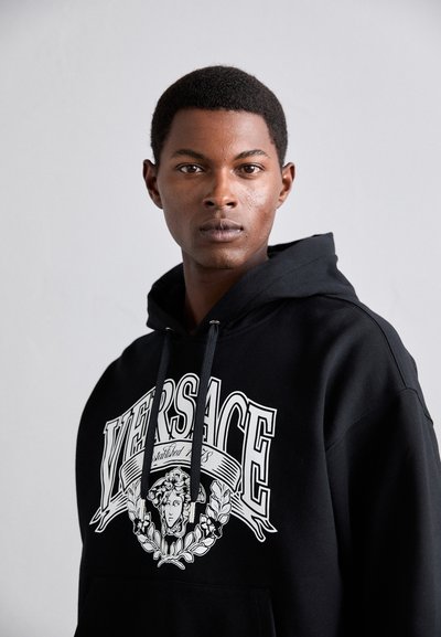 Giovane uomo con capelli corti che indossa una felpa nera di Versace con il logo di Medusa, che guarda direttamente davanti a sé su uno sfondo chiaro e semplice.