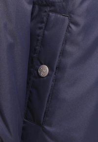 Granatowy materiał z pionową kieszenią zapinaną na metalowy guzik z wygrawerowanym stylizowanym logo pszczoły.